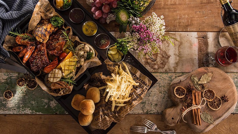 Une jolie planche remplie de viandes, de frites, de pains et de sauces posée sur une table en bois.